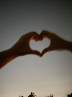 two hands making a heart shape with their fingers at night sky and trees in the background