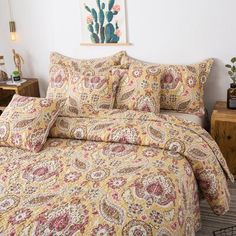 a bed covered in a yellow paisley comforter next to a night stand with a painting on the wall