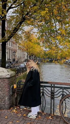 a woman standing on the side of a river next to a tree filled with leaves