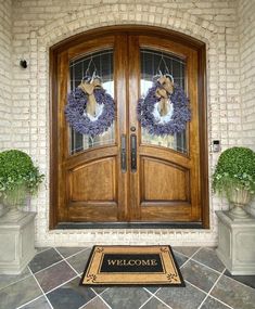 two wreaths on the front door of a house with welcome mat and potted plants