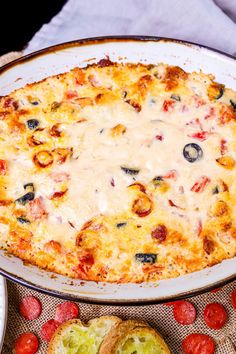 an oval casserole dish with cheese and olives on it next to bread