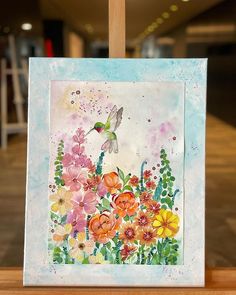 a painting with flowers and a hummingbird in the sky on a wooden easel