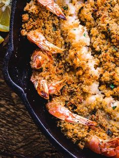 a skillet filled with shrimp and crumbs next to lemon wedges