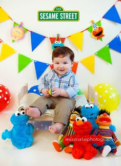 a baby sitting on a chair surrounded by stuffed animals and sesame street sign in the background