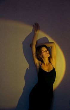 a woman in a black dress is leaning against a white wall with her arms up