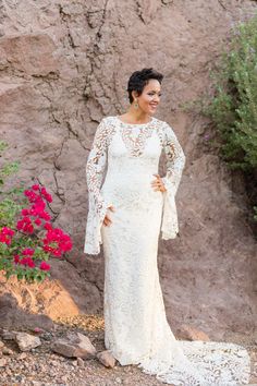 a woman standing in front of a rock wall wearing a white lace dress with bell sleeves