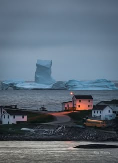 an iceberg floating in the ocean next to small houses and buildings at dusk with lights on