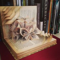 an open book with origami flowers on it sitting next to bookshelves