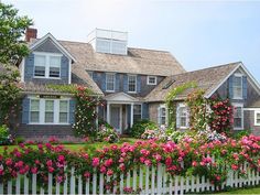 a white picket fence with pink flowers in front of a large gray house and trees