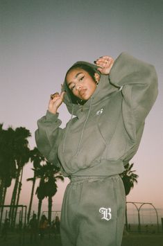 a woman wearing a hoodie and sweatpants poses for a photo in front of palm trees