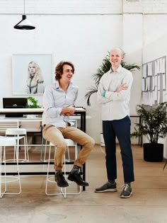 two men standing in an office with their arms crossed and one sitting on a chair