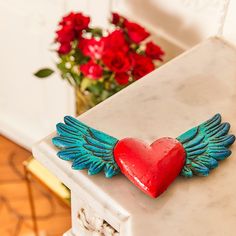 a red heart with wings sitting on top of a white table next to some flowers