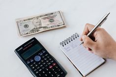 a person writing on a notepad next to a calculator, pen and money