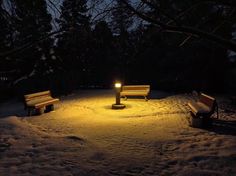 two park benches sitting in the middle of a snow covered field at night with a light shining on them