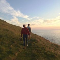 two people walking up a grassy hill towards the ocean at sunset or sunrise with one person carrying a backpack