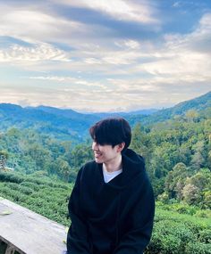 a young man sitting on a bench in front of a scenic mountain valley with tea bushes