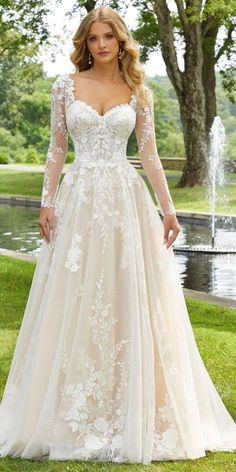 a woman wearing a wedding dress with long sleeves and flowers on the skirt is standing in front of a pond