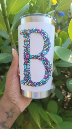 a hand holding a cup with beaded initial b on the front and bottom, in front of green leaves