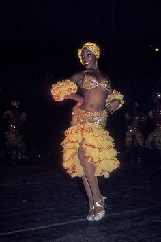 a woman in a yellow skirt and head wrap dancing on stage with other dancers behind her