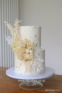 a three tiered wedding cake with white and gold flowers on the top, sitting on a wooden table