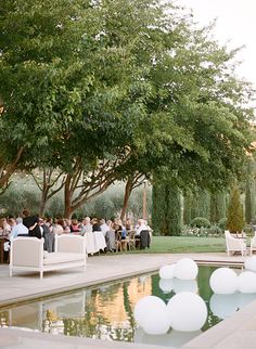 a group of people sitting at tables next to a pool with white balloons floating in the air