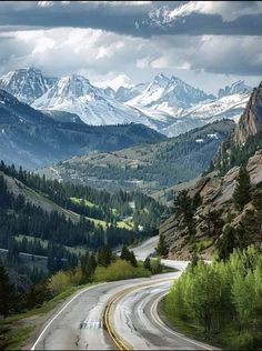 an empty road in the mountains with snow capped mountains on either side and trees lining both sides