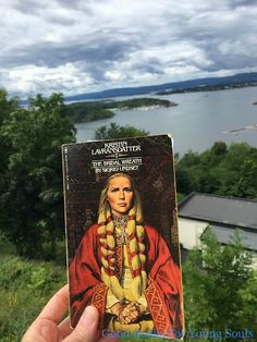 a person holding up a book in front of a lake and mountains with clouds overhead