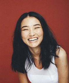 a woman smiling with her eyes closed in front of a red wall and wearing a white tank top