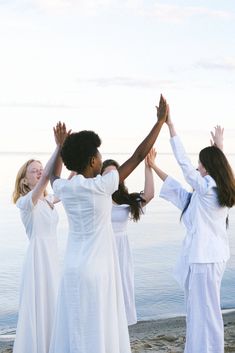 four women in white dresses standing on the beach with their hands up to each other