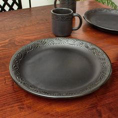 a wooden table with two black plates and cups on it, along with a plant in the background