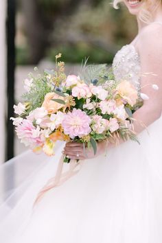 a woman in a wedding dress holding a bouquet of pink and yellow flowers with greenery