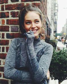 a woman standing in front of a brick wall with her hands on her face and looking at the camera