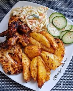 a white plate topped with chicken wings, potatoes and cucumbers next to coleslaw