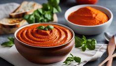 a bowl of carrot hummus next to two bowls of bread and fresh basil leaves