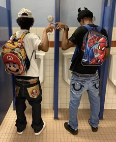 two boys with backpacks standing in front of urinals and one boy is holding an object