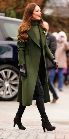 a woman in green coat and black boots walking down the street with her hand on her hip