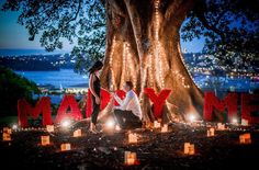 two people sitting on the ground in front of a tree with lit up letters and candles