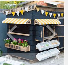 an outdoor food stand with flowers and bunting on the roof, in front of a blue building