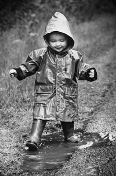 a little boy that is standing in the water wearing a rain coat and boots with his hands out