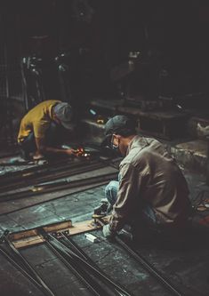 two men working on some metal in a factory or repair area, one is bending over and the other is kneeling down