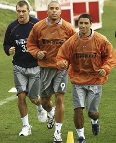 two men running on a field with one man wearing an orange shirt and the other in grey shorts