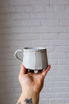 a hand holding a white ceramic cup in front of a brick wall with an arrow tattoo on it