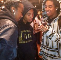 three young people looking at something on their cell phone while standing in a crowded room