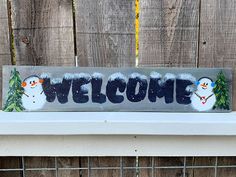 a welcome sign with snowmen and pine trees painted on the side of a wooden fence