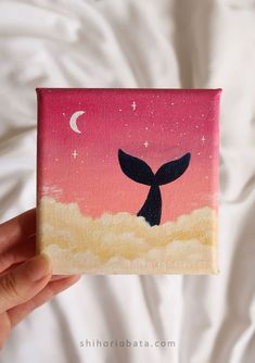 a hand holding up a small painting of a whale tail in front of a pink sky