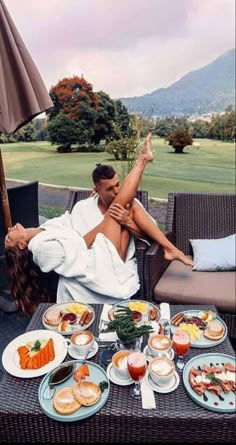 a man and woman sitting on a patio with food in front of them, under an umbrella