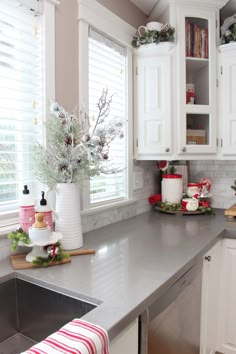 a kitchen with white cabinets and silver counter tops, decorated for christmas time in front of the window