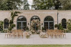 an outdoor ceremony setup with wooden chairs and greenery on the side of the building