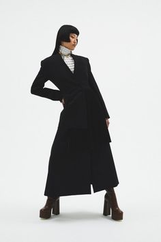 a woman in a long black coat and boots standing with her hands on her hips