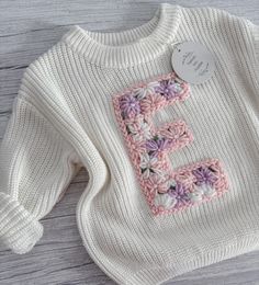 a white sweater with pink and purple crocheted letters on the front, sitting on a wooden surface